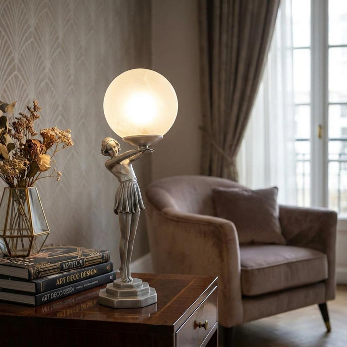 Art Deco silver figurine table lamp holding a glowing orb on a wooden table in a living room.