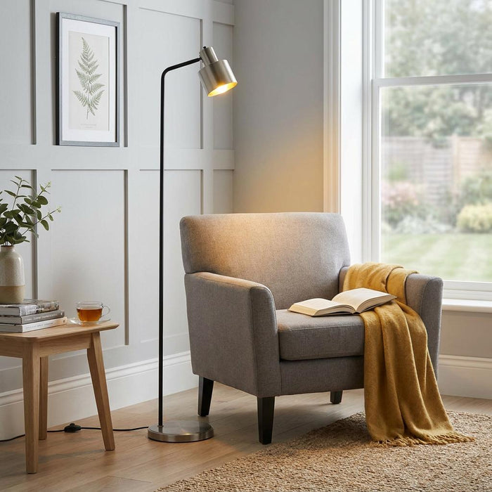 Modern floor lamp with a black stem and brushed silver head casting a warm glow over a grey armchair.