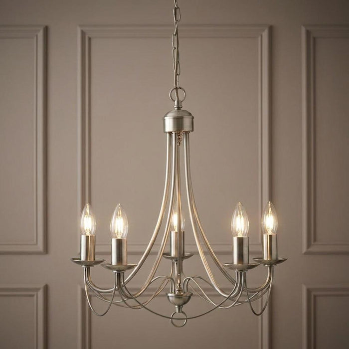 Five-light antique brass pendant chandelier with lit candle bulbs against a decorative panelled wall.