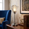 Chrome table lamp with a ribbed glass shade sitting on a wooden table beside a plush blue armchair.
