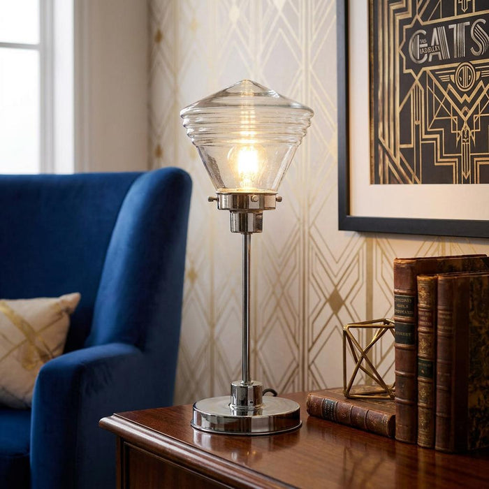 Chrome table lamp with a ribbed glass shade sitting on a wooden table beside a plush blue armchair.