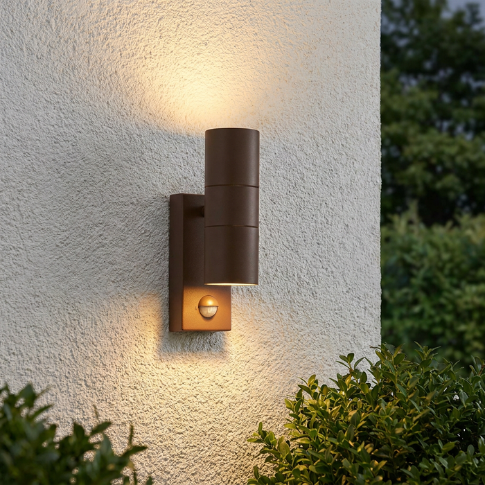 Brown cylindrical up and down outdoor wall light with PIR sensor illuminating a white textured wall.