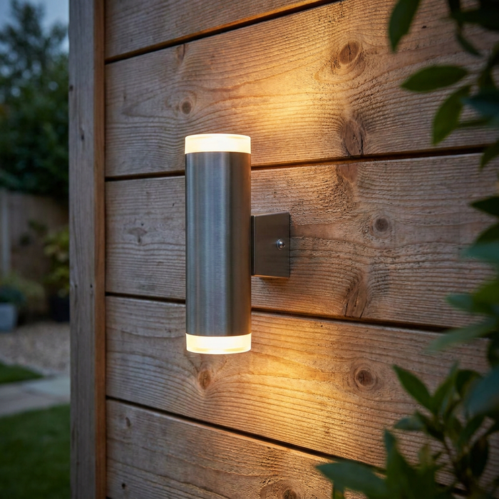 Satin silver cylindrical up and down outdoor wall light mounted on a wooden fence with a warm glow.