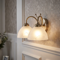 Milanese two-light brass wall sconce with frosted alabaster glass shades illuminated on a patterned wall.