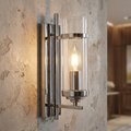 A sleek chrome wall sconce with a cylindrical glass shade and glowing candle bulb on a textured wall.
