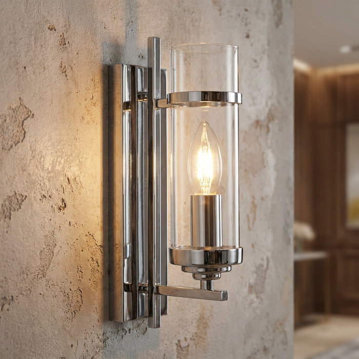 A sleek chrome wall sconce with a cylindrical glass shade and glowing candle bulb on a textured wall.