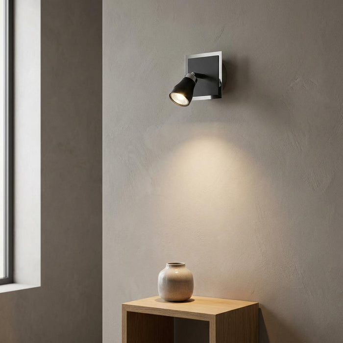 Milo black and chrome single spotlight mounted on a textured wall, illuminating a vase on a wooden shelf.