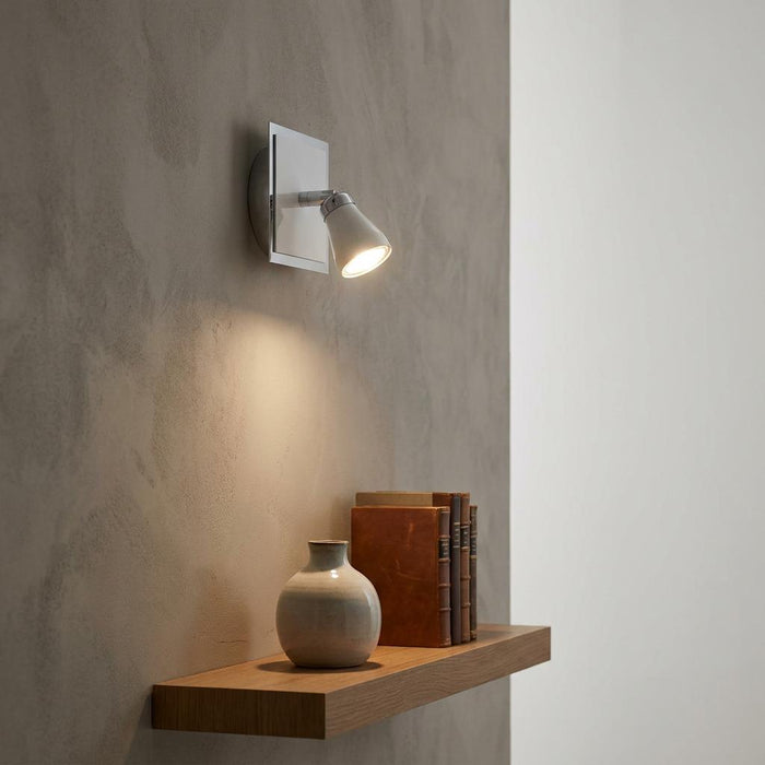 Milo white and chrome spotlight illuminating a ceramic vase and books on a wooden shelf.