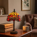 Orange Tiffany dragonfly table lamp glowing on a wooden side table next to vintage books and a steaming mug.
