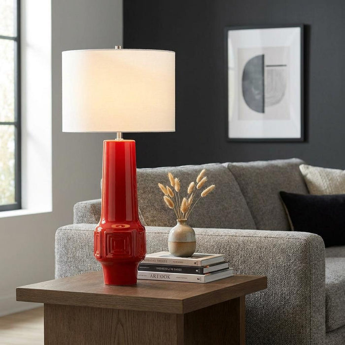 Red ceramic table lamp with a white drum shade on a wooden side table in a modern living room setting.