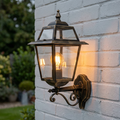 Traditional black and gold lantern wall light with a lit bulb mounted on a white brick wall.