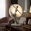 Silver Art Deco figurine lamp of a dancing woman against a glowing circular glass backdrop on a polished wooden table.