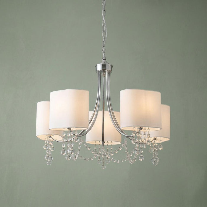 Chrome and crystal pendant light with five lit white shades and hanging glass droplets against a green wall.
