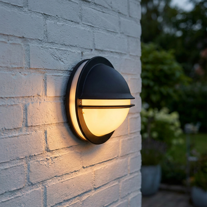 Black eyelid-style outdoor wall light glowing on a white brick wall in a garden setting.