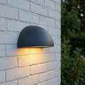 Norlys Halden graphite dome-shaped outdoor wall light casting a warm glow on a white brick wall.