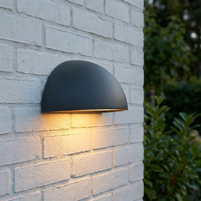 Norlys Halden graphite dome-shaped outdoor wall light casting a warm glow on a white brick wall.