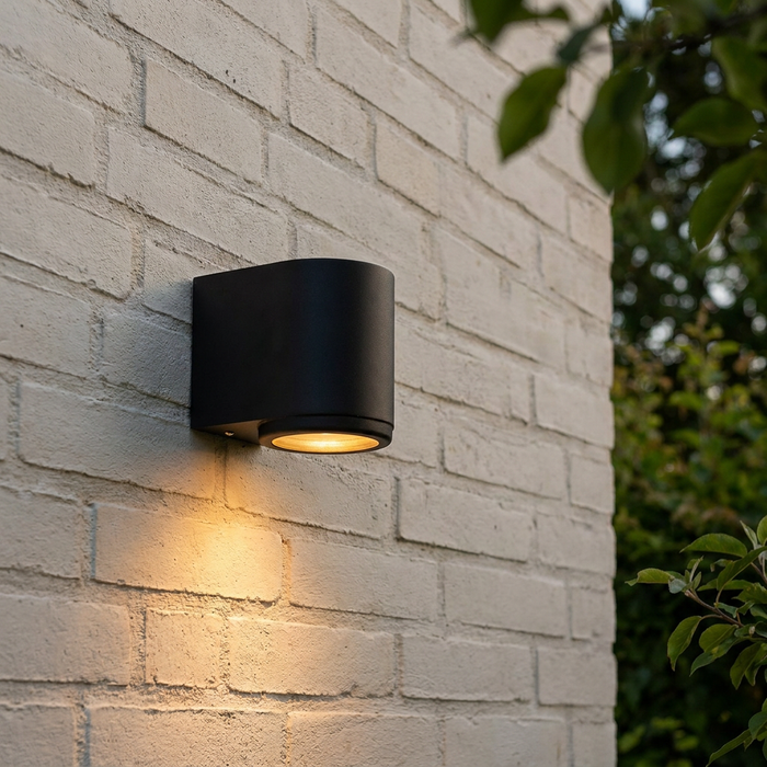 Sleek black outdoor wall light casting a warm downward glow on a white brick wall.