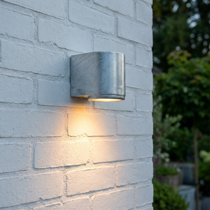 Galvanized steel wall light mounted on white brick, casting a warm downward glow.