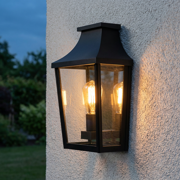 Black lantern outdoor wall light with an Edison bulb, glowing warmly on a textured white wall.