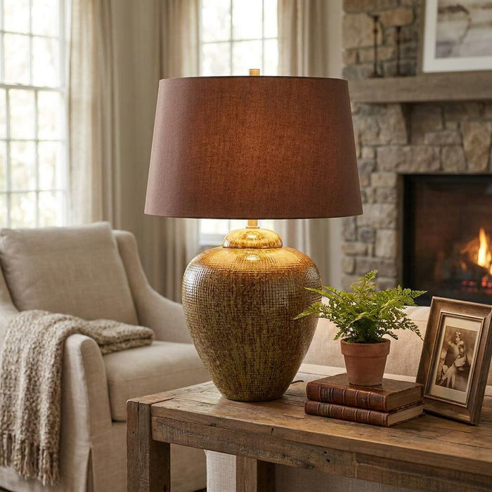 Green and brown textured ceramic table lamp with a dark brown shade on a wooden table in a cozy living room.