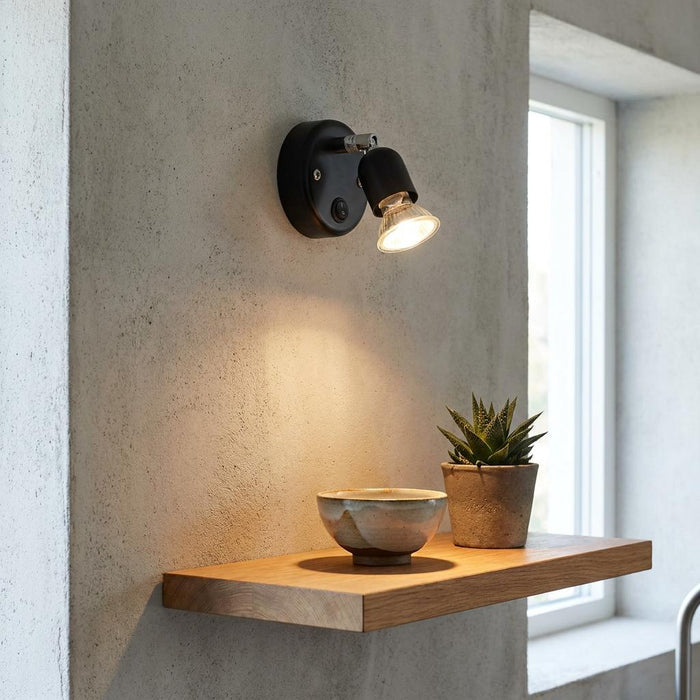 Black single switched spotlight mounted on a textured wall, illuminating a bowl on a wooden shelf.