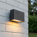 Dark grey rectangular LED wall light mounted on a white brick wall, emitting a warm downward glow.