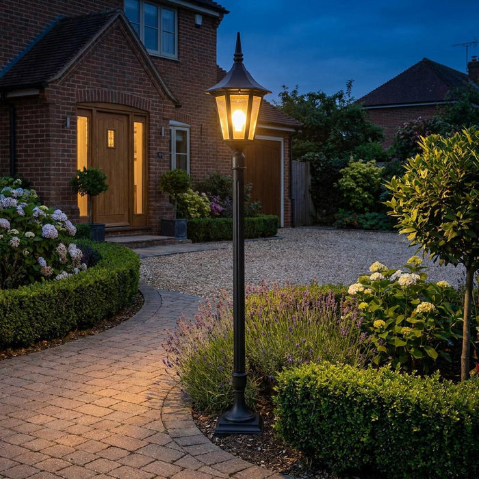 Black outdoor mini lamp post glowing beside a brick path and lush garden at twilight.