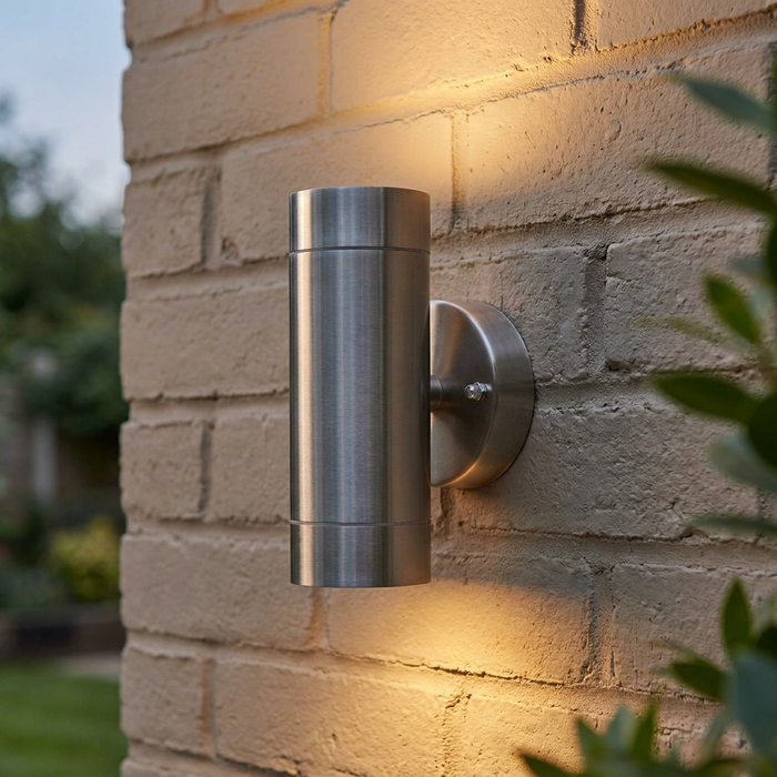 Cylindrical stainless steel up and down light mounted on an exterior brick wall, casting a warm glow.