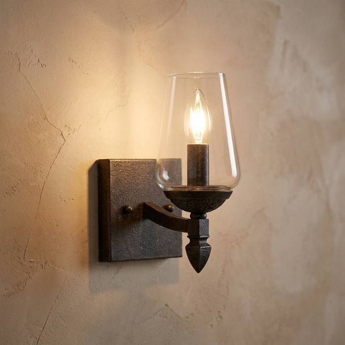 Single wall light with a clear tapered glass shade and dark textured finish glowing against a neutral plaster wall.