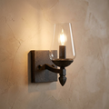 Single wall light with a clear tapered glass shade and dark textured finish glowing against a neutral plaster wall.