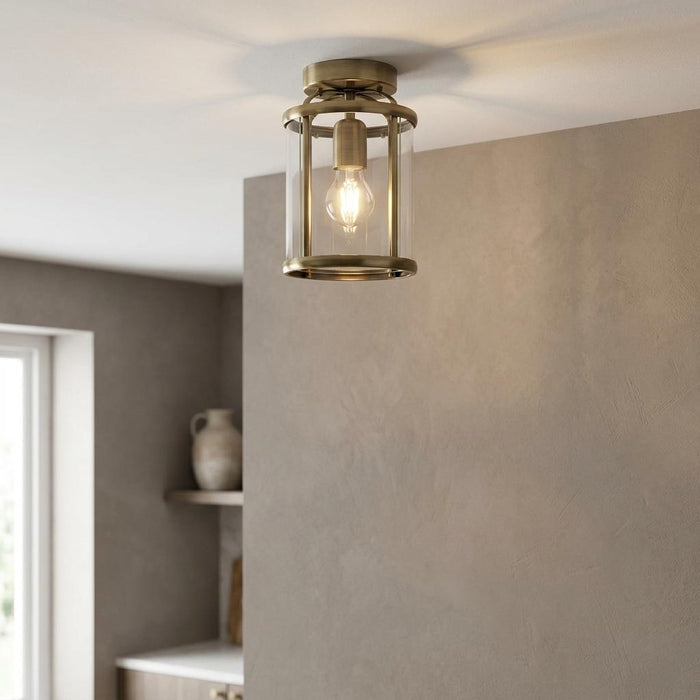 Antique brass flush ceiling light with a clear glass cylinder shade illuminated in a neutral interior.