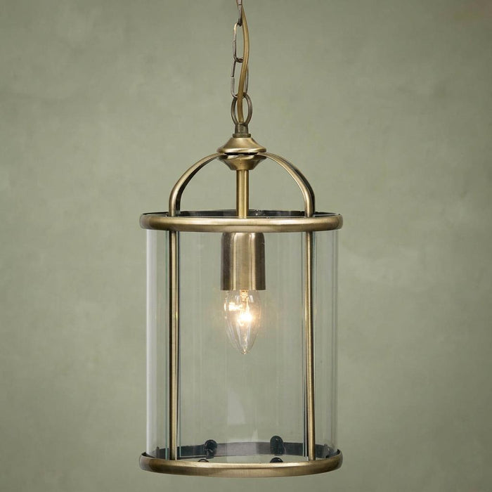 Antique brass ceiling lantern with a cylindrical glass shade and a glowing bulb against a green wall.