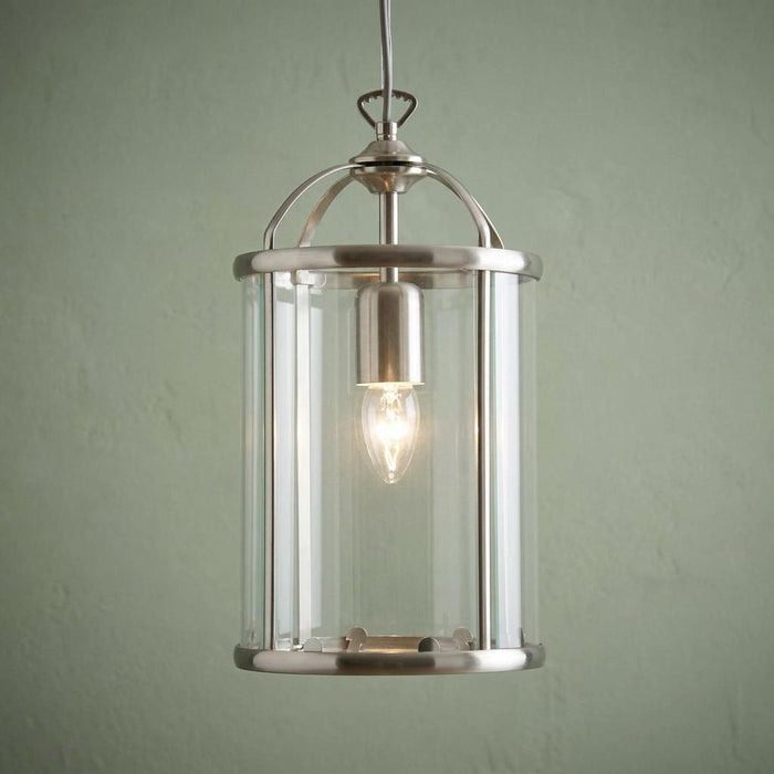 Antique chrome cylindrical glass lantern with a single lit bulb hanging against a pale green wall.