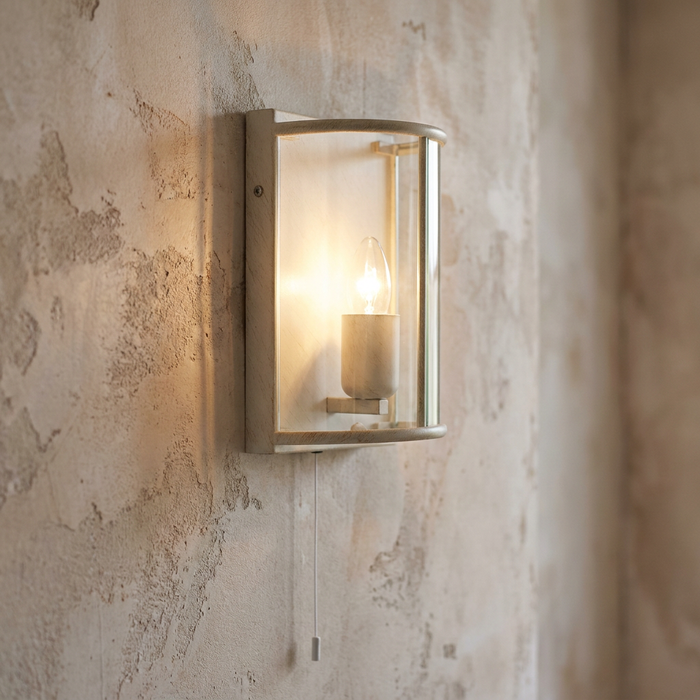 A cream gold finish wall light with curved glass and pull cord glowing on a textured rustic wall.