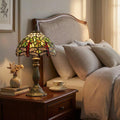 Green dragonfly Tiffany bedside lamp glowing on a wooden nightstand next to a bed with a beige headboard.