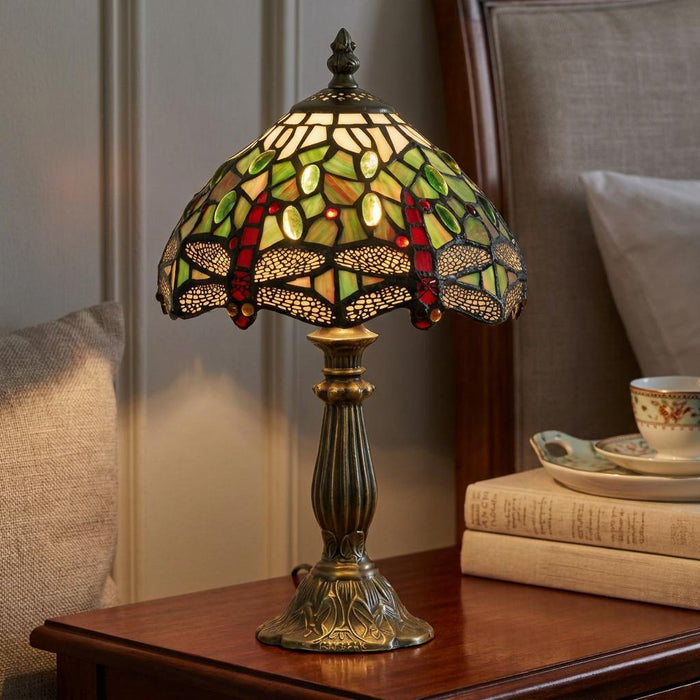 A green Tiffany dragonfly lamp with a stained glass shade on a bedside table beside books.
