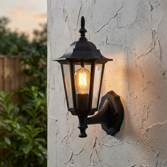 Traditional black lantern wall light with a warm filament bulb mounted on a textured white exterior wall.