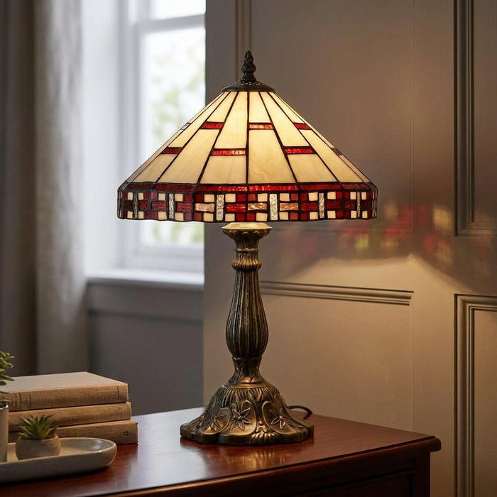 Tiffany-style table lamp with a cream and red geometric glass shade on an ornate bronze base, lit on a wooden desk.