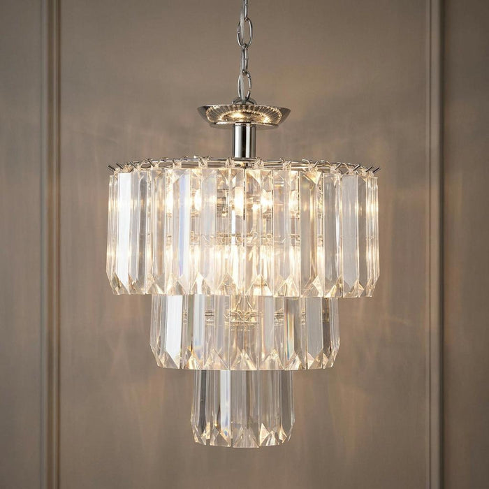 Three-tier clear acrylic and chrome pendant light with glowing bulbs hanging against a beige paneled wall.