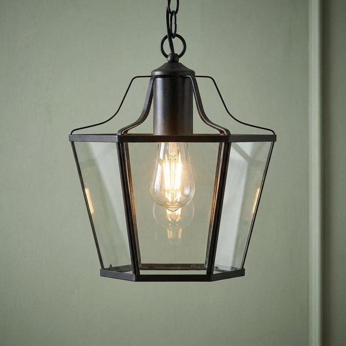 Black lantern-style pendant light with clear glass panels and an Edison bulb against a green wall.