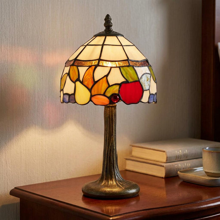 Warmly lit fruit-patterned Tiffany lamp on a bronze base sitting on a wooden nightstand beside stacked books.