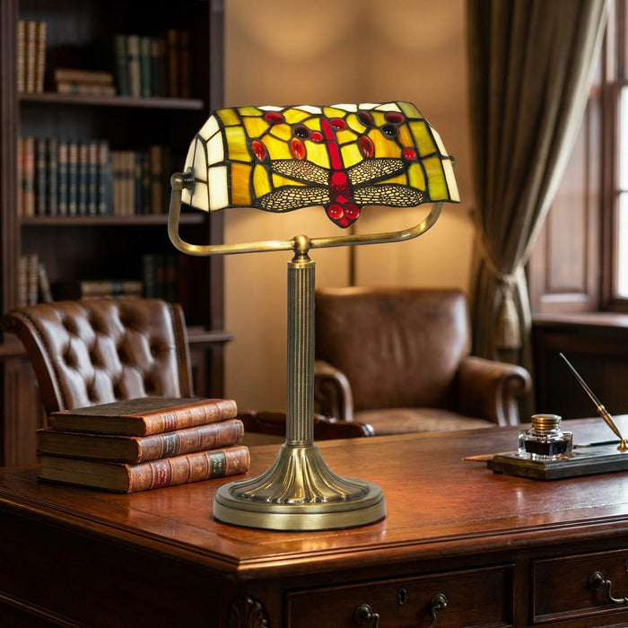 Stained glass dragonfly bankers lamp with red and green details on a vintage desk in a library.