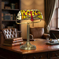 Stained glass dragonfly bankers lamp with red and green details on a vintage desk in a library.