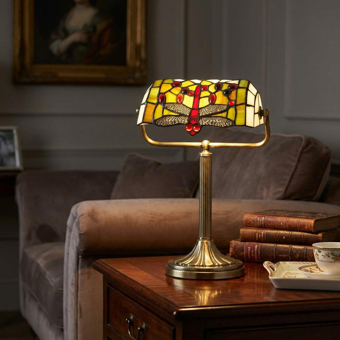 Tiffany style dragonfly banker's lamp with a stained glass shade on a wooden table beside a stack of vintage books.