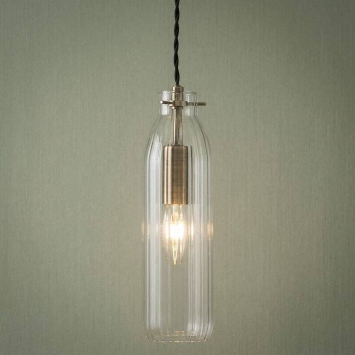 Antique brass pendant light with a fluted clear glass bottle-shaped shade against a green wall.
