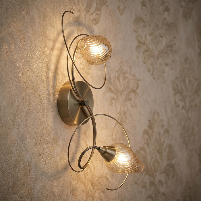 Lugh twin antique brass wall light with swirling metal arms and ribbed glass shades on damask wallpaper.
