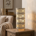 Cylindrical ivory filigree table lamp with crystal accents glowing on a wooden side table in a cozy living room.