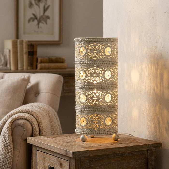 Cylindrical ivory filigree table lamp with crystal accents glowing on a wooden side table in a cozy living room.