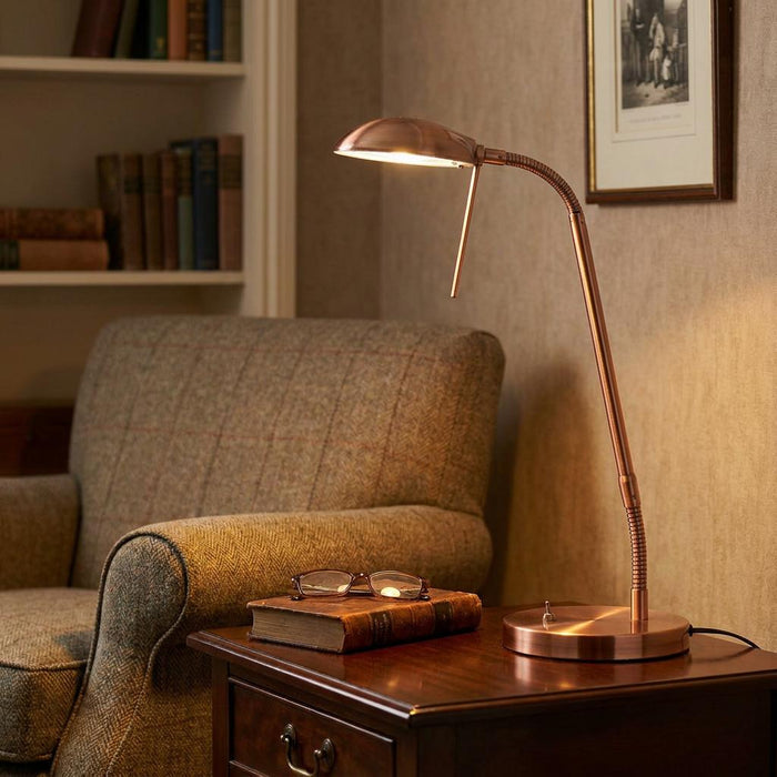Copper flexible head table lamp illuminating a book and glasses on a wooden side table next to an armchair.