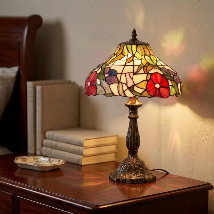 Ornate Tiffany table lamp with a floral peony stained glass shade glowing on a dark wood bedside table.